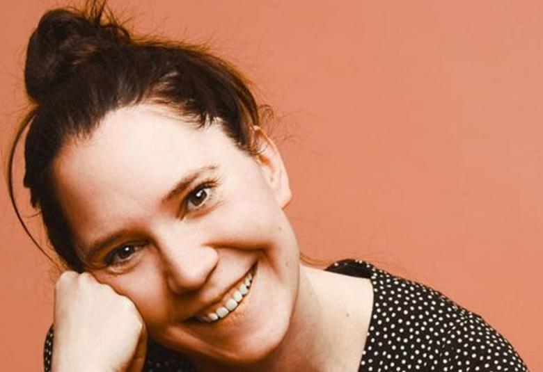 Kuvassa on Jenna Kostet kasvokuvassa Hänen kasvonsa ovat vinottain ja hän nojaa oikealla kädellään poskeensa. Kostetilla on tummat hiukset nutturalla. Tausta on vaalean ruskea.