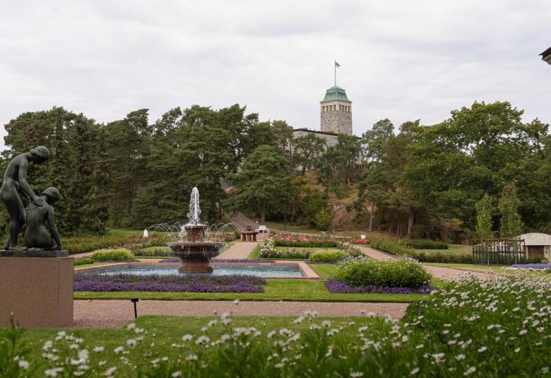 Kuvassa on Kultarannan puutarhan maisemaa ja etualalla on vasemmalla tumma patsas sekä muotopuutarhaa. Välissä on lehtipuustoa ja keskellä on torni. 