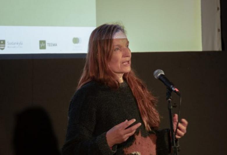Kuvassa on Johanna Vuoksenmaa puolivartalokuvassa pitämässä esitelmää. Hän on puhumassa vinottain oikealle. Taustalla on projektorin valoa ja vaalean vihreää seinää. Osa valosta menee hänen otsansa yli. Hänellä on tumma pusero päällään.