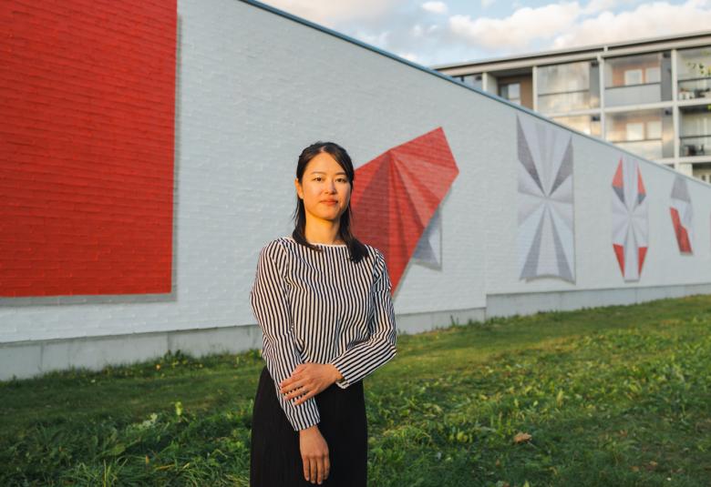 Kuvassa on pitkää seinämää ja siinä on vasemmala on maalattu punainen iso neilö. Vaalealla seinämällä on punainen origami sekä neljä muuta kuviota, joissa on myös harmaata. Edessä on seisomassa tekijä Momo Yoshino.