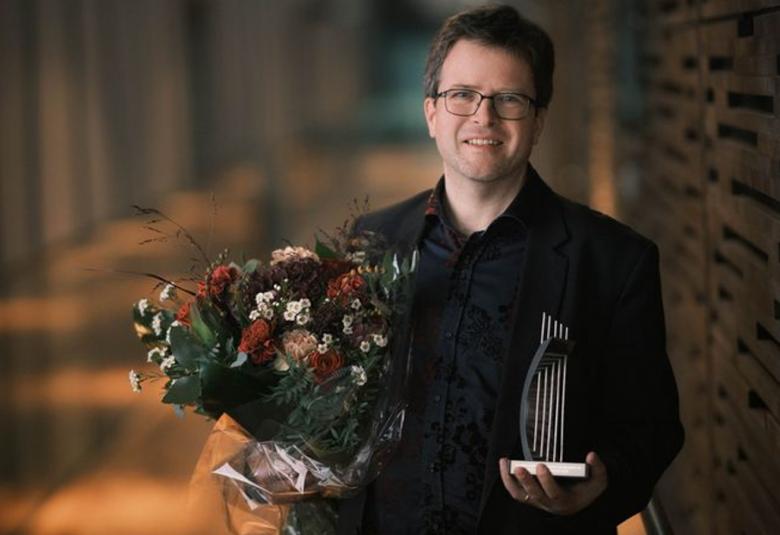 Kuvassa on Nils Scweckendiek puolivartalokuvassa ja hänellä on musta takki päällään. Vasemmassa kädessään hänellä on kukkakimppu ja oikeassa kädessään palkintoveistos, jossa on kaari ja nuottirivistö ylöspäin.