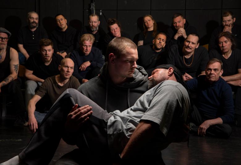 Kuvassa etualalla mies kantaa nukahtanut nuorempaa miestä. Taustalla on joukko vankeja, jotka katsovat miehiin. Kuvan sävy on tumma.