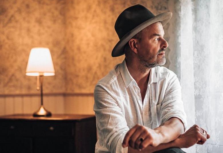 Kuvassa on oikealla Tuure Kilpeläinen istumassa ja hänellä on valkoinen paita ja harmaa hattu. Hän katsoo oikealle ja on sivuttain. Taustalla näkyy vaalean ruskeaa seinää, jossa on kulma. Seinän vieressä on ruskea matala hyllykkö ja sen päällä on pöytälamppu valaistuna.