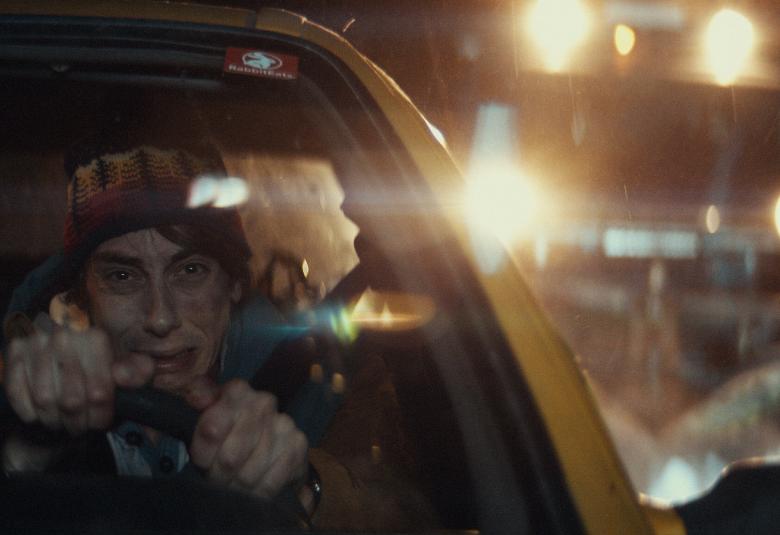 Kuvassa on vasemmalla nuori mies ajamassa autoa yöllä ja autoa näkyy puoliksi. Taustalla näkyy valoja öisellä tiellä ja kuorma-auton valot.