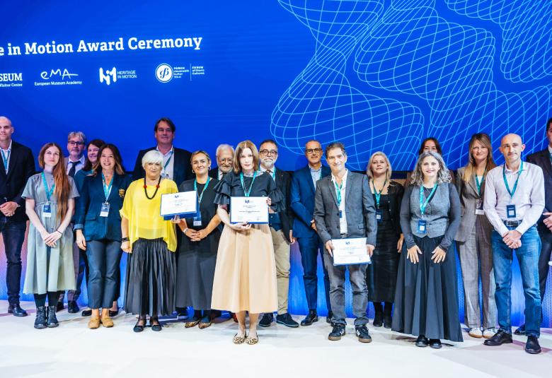 Kuvassa on iso joukko ihmisiä lavalla ja keskellä edessä on kaksi naista sekä yksi mies, jotka pitävät valkoista kunniakirjaa kädessään. Tausta on kirkkaan sininen ja valkoisella lukee Heritage in Motion Award Academy.