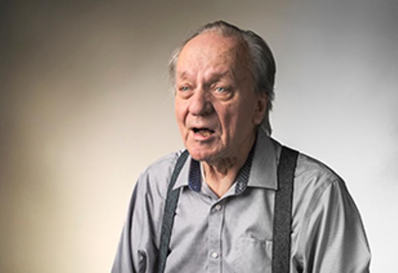 Kuvassa on Esko Roine istumassa. Hänellä on harmaa paita päällään ja tumman harmaat henkselit. Tausta on ruskehtava.