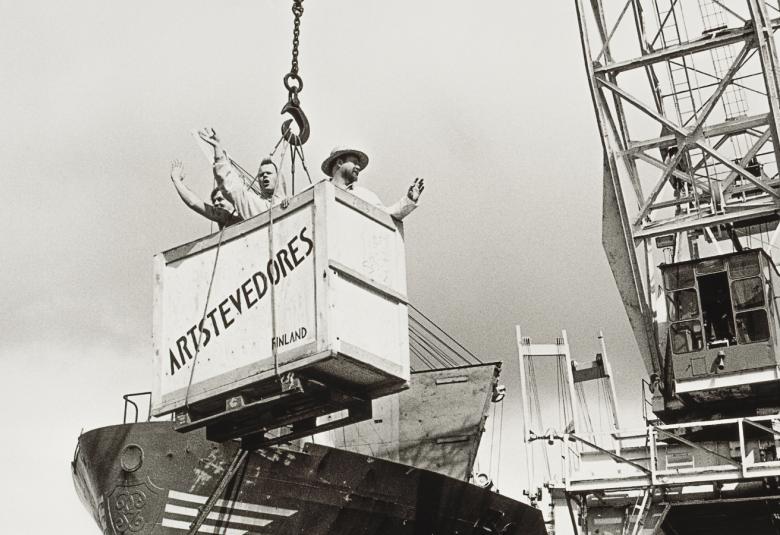 Kuvassa on satamanosturi laskemassa puulaatikkoa laivan kannelle. Laatikossa on teksti Artstevendores.  Kuva on musta-valkoinen.