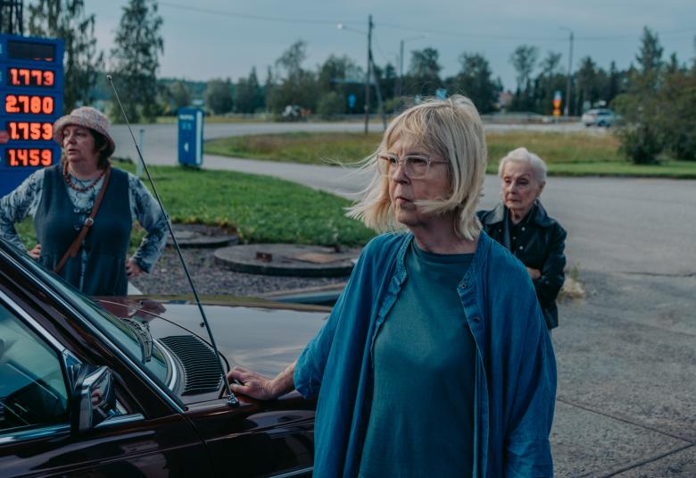 Kuvassa on tumman auton keula huoltoasemalla. Taustalla näkyy sininen polttoaineen hintataulu hintoineen. Auton edessä seisoo Leena Uotila sinisessä villatakissa ja mekkossa. Takana on Sanna Palo sinisessä liivimekossa. ja Uotilan takana seisoo Seela Sella. Taustalla näkyy nurmikenttiä ja sisääntulotiet.