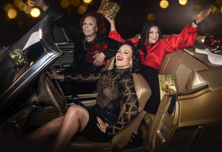 Kuvassa on etualalla Erja Lyytinen istumassa avoauton ruskealla istuimella ja hänen vieressään Kärtsy Hatakka. Takapenkillä istuu Hanna Pakarinen punaisessa puserossa molemmat kädet levitettynä ylöspäin. 