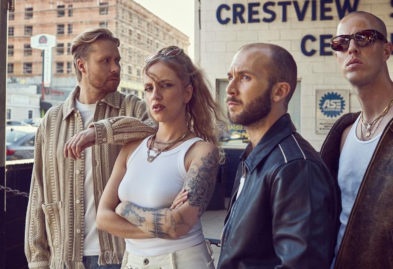 Kuvassa on Haloo Helsingin jäsenet seisomassa tilassa, jossa lukee seinässä Crestview Center. Vasemmalla on mies vaalean ruskeassa villatakissa vieressäänElli valkoisessa hihattomassa t-paidassa. Oikealla on kaksi miestä ja heillä on tummat takit päällään. Lasi-ikkunan takaa näkyy vaalean ruskea iso kerrostalo.