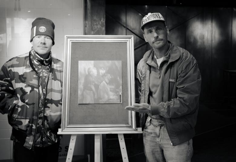 Kuvassa ovat vasemmalla VilleGalle ja oikealla Jare. Heidän välissään on raamitettu taulu varustettuna kolmella jalalla. Kuva on musta-valkoinen.