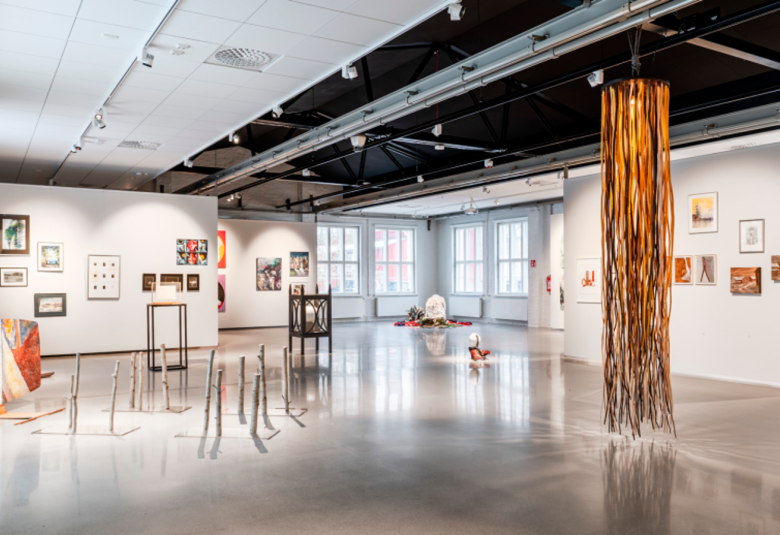 Kuvassa on vaalean harmaat seinät taidenäyttelytilassa ja siinä on eri kokoisia taideteoksia seinillä ja takana yksi pylväs.