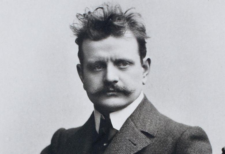 Kuvassa on  Jean Sibelius puolivartalokuvassa ja hänellä on hiukset päälaella pörhöllään ja paksu takki päällään sekä valkoiset kaulukset. Kuva on musta-valkoinen.