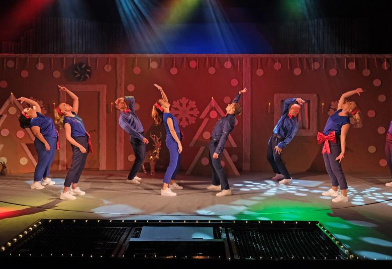 Kuvassa on kahdeksan henkilöä sinisissä asuissa taipuneina taaksepäin toinen käsi ojennettuna ylös. Taustalla on ruskeaa seinämää, jossa on ruskehtavia joulukuusia.