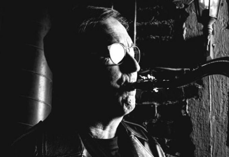 Kuvassa on Jussi Saksa soittamassa saksofonia puolivartalokuvassa. Kuva on tumma ja silmälasit kiiltävät ja kasvot näkyvät osittain.