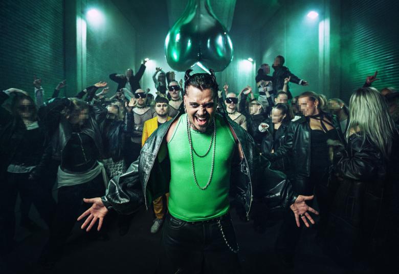 Kuvassa on keskellä Käärijä vihreässä puserossa ja mustassa nahkatakissa. Taustalla on joukko ihmisiä ja valoja ohjattuna heihin. 