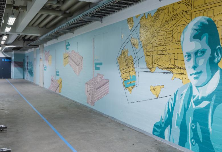 Kuvassa on käytävä, jossa on oikean puoleinen seinä ja sen pohja on vaalean sininen.  Seinällä on karttapohjamaisia kuvioita ja oikealla keltainen niemi.  Lähinnä oikealla on iso miehen rintakuva vaalean sinisessä takissa.