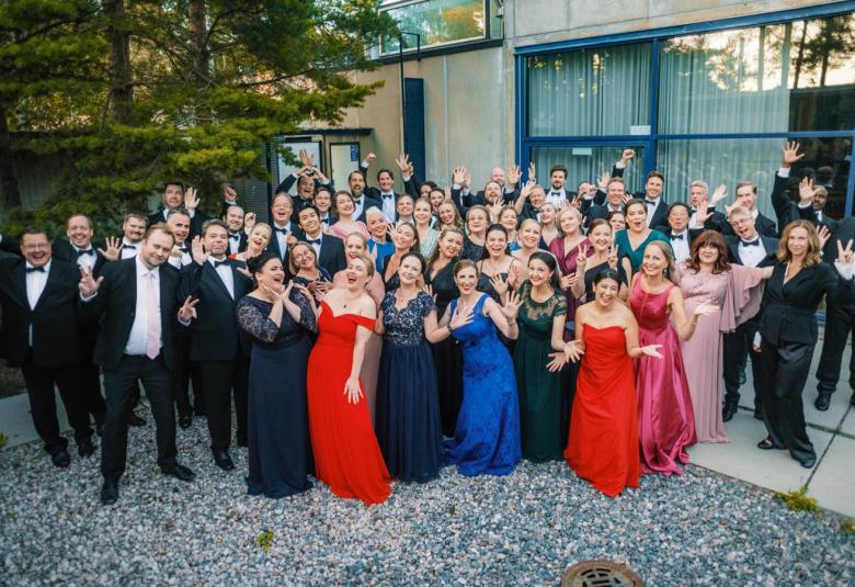 Kuvassa on koko kansallisoopperan kuoro seisomassa pihalla. Vasemmalla ja takana ovat miehet musta-valkoisissa puvuissa. Edessä ovat naiset pitkissä, eri värisissä pitkissä iltapuvuissa. 