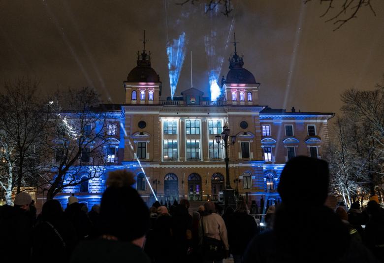 Kuvassa on Oulun kaupungintalo yöllä ja sitä valaistaan sinisillä valoilla molemmilta reunoilta. Talossa on kaksi tornia ja kolme korkeaa valaistua ikkunaa.
