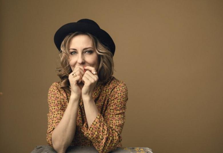 Kuvassa on Mariska puolivartalokuvassa ja hänellä on musta hattu päässään sekä kellertävä, pikkukuvioinen pusero päällään. Hänellä on molemmat kädet kevyesti nyrkissä suun edessä. Tausta on ruskehtavaa seinää.