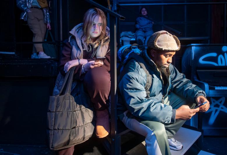 Kuvassa on oikealla nuori mies istumassa karvahatussa ja toppatakissa. Hänen takanaa vasemmalla on nuori nainen, jonka hiukset roikkuva ja hän nojaa vasempaan jalkaansa. Tausta on osittain tumma. 