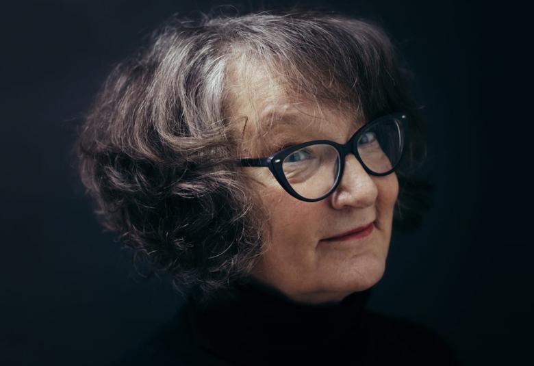 Kuvassa on Monika Fagerholmin kasvokuva ja hänellä on kihartavat tumman harmaat hiukset  sekä mustasankaiset silmälasit. Tausta on musta.