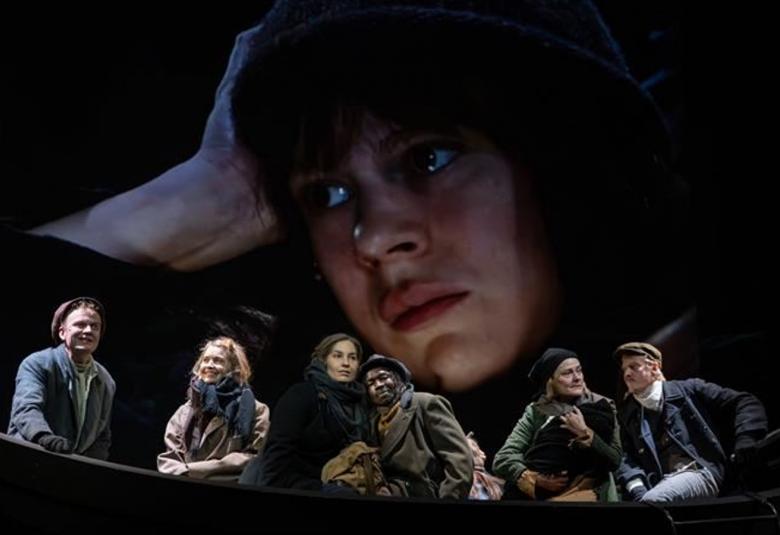 Kuvassa on veneessä kuusi henkilöä tummahkoissa vaatteissa. Taustalla on tummalla taustalla nuoren naisen kasvokuva lierihatussa.