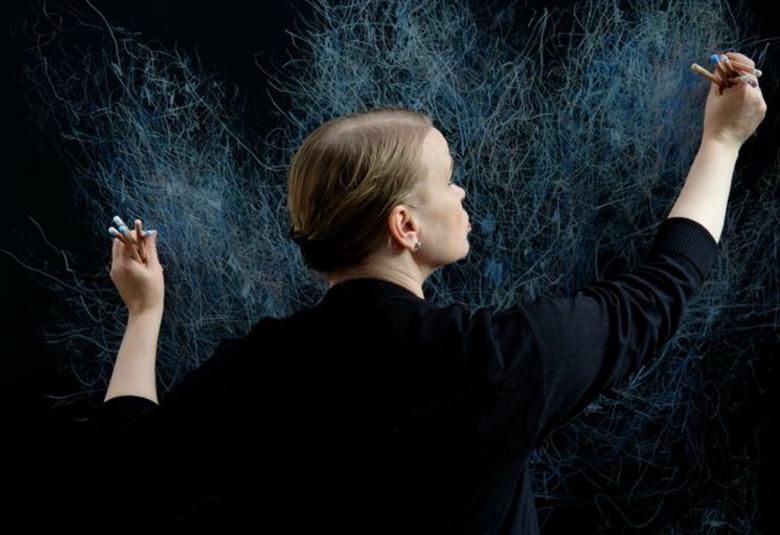  Kuvassa on Varpu Rossi piirtämässä tummalle seinälle puolivartalokuvassa kädet levitettyinä maalausasentoon. Hänellä on musta pusero.