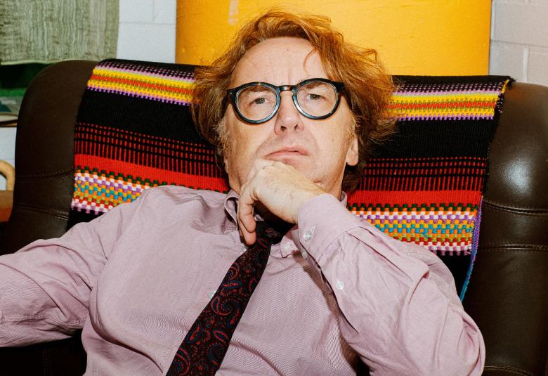 Kuvassa on Pertti Jarla istumassa nojatuolissa puolivartalokuvassa. Hänellä on lilahtava paita ja tumma kravatti. Selkänojassa on tumman ja punaisen raidallinen matto. 