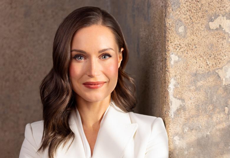 Kuvassa on Sanna Marin puolivartalokuvassa ja hänellä on valkoinen jakku päällään, jossa on v-aukko. Hänellä on pitkät tummat hiukset. Tausta on vaalean ruskeaa kiviseinää.