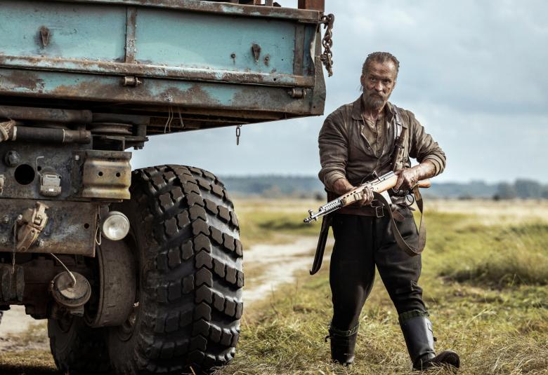 Kuvassa on vasemmalla kuorma-auton korkea takaosa.  Sen vieressä seisoo Jorma Tommila ase kädessään vihreässä maastopuvussa. Taustalla on vihreää ruohoa ja hiekkatietä. 