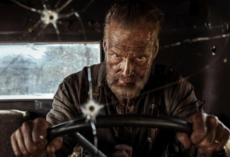 Kuvassa on Jorma Tommila ajamassa sotilasajoneuvoa ja tuulilasissa on luodin reikä.  Autossa on tummat seinät ja takaa näkyy avoin ikkuna.