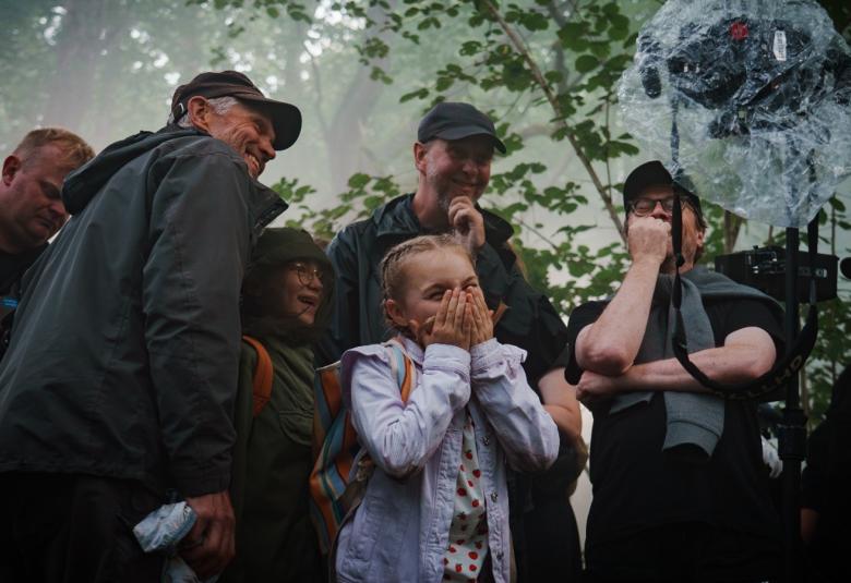 Kuvassa on Siiri Virtanen keskellä vaalean sinisessä takissa. Ympärillä on aikuisia miehiä ja naisia tummissa takissa ulkona lehtipuiden alla. 