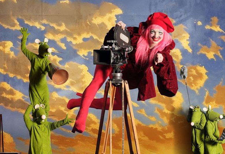 Kuvassa taustalla kelta-harmaita pilviä sinisellä taivaalla. Vasemmalla on kaksi vihreätä hiireksi pukeutunutta hahmo ja toinen seisoo okapäillä. Keskellä on vanhanaikainen filmikamera ja siinä päällä roikkuu nainen punaisissa saappaissa legginsseissä sekä viininpunaisessa takissa. Takana on pieninä kaksi vihreätä hiirtä.