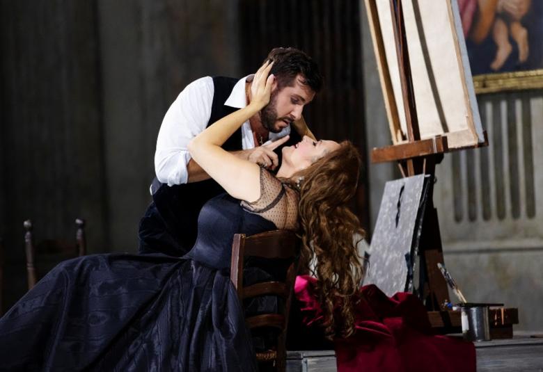 Kuvassa on Toscan roolin laulaja tumman sinisessä pitkässä puvussa istumassa tuolissa ja nojaa selkänojaan.  Hänen ylläpuolellaan on kuvartuneena kasvoja kohti mies valkoisessa puserossa ja mustassa liivissä. Tausta on tumma.