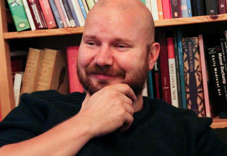 Kuvassa on Tuomo Rämö puolivartalokuvassa istumassa takanaa kirjahyllyä. Hänellä on musta paita ja hän pitää oikeaa kättä leuassaan.