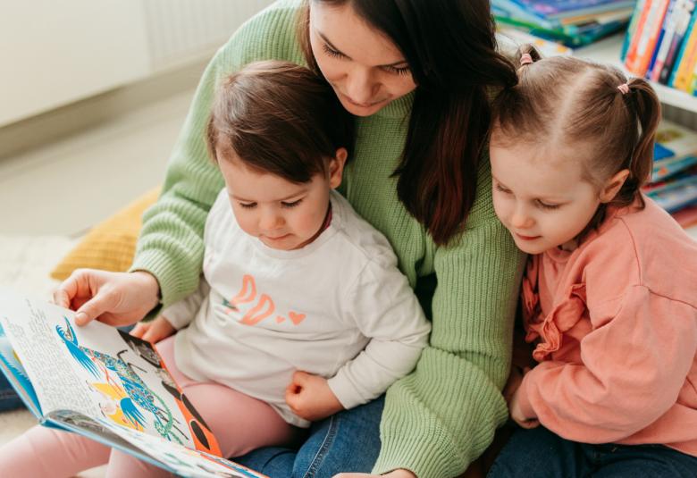 Kuvassa on nuori nainen, joka lukee kirjaa kahdelle sylissään olevalle lapselle. He ovat noin neljä- ja kuusivuotiaita. 