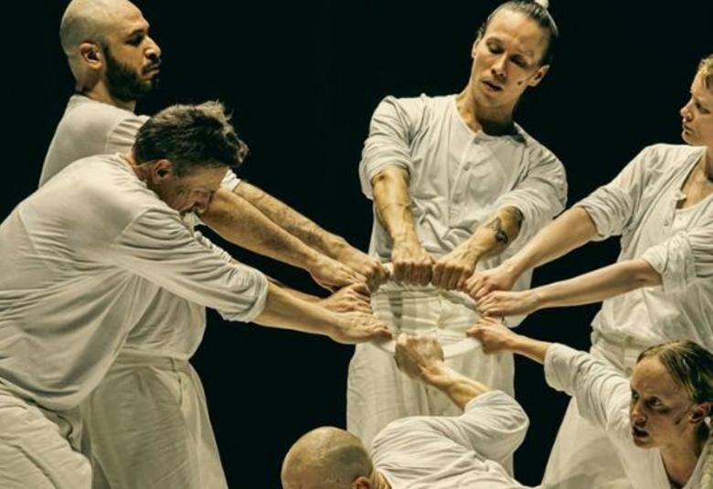 Kuvassa on viisi miestanssijaa ja yksi näinen vaaleissa kangasasuissa ja he pitävät kahdella kädellä kiinni vaaleasta renkaasta. Edessä oleva tanssija on alhaalla kumartuneena.