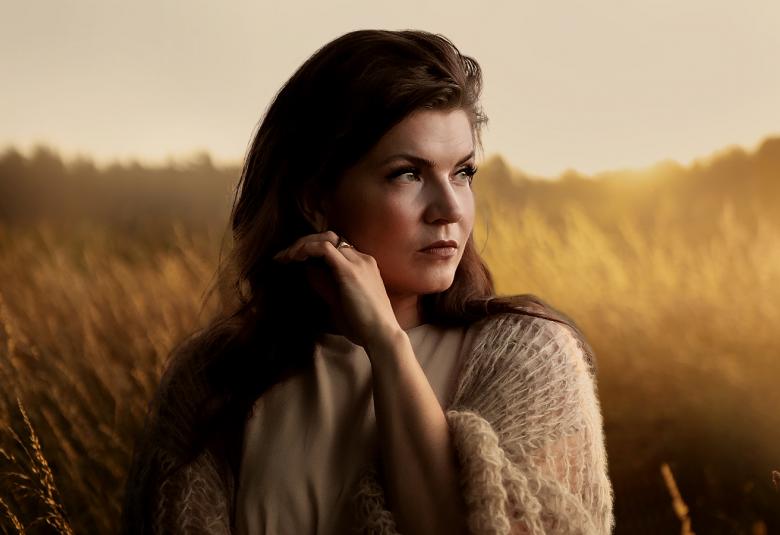 Kuvassa on Suvi Teräsniska puolivartalokuvassa ja hän katsoo oikealle. Taustalla on auringonlaskun kellertävää ja viljapelto, jossa on usvaa.