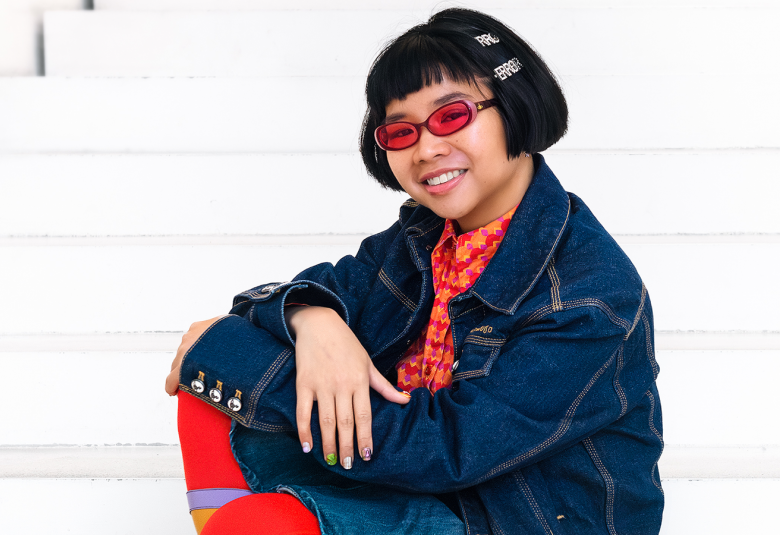 Kuvassa on valkoisilla portailla istumassa Natasha Tontey sinisessä farkkutakissa ja punaisissa sukissa.