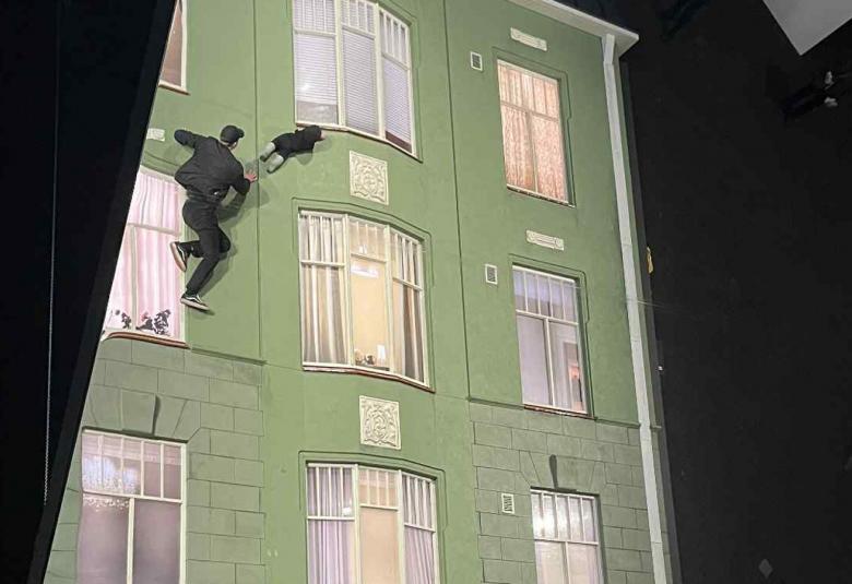 Kuvassa on molemmin puolin mustaa ja keskellä on vaalean vihreä vanha kerrostalo, jossa on kolmeen osaan jakautuneet ikkunoita kolmessa rivissä. Vasemmalla ylhäällä on nuori mies mustassa puvussa mukanaan vauva kiipeämässä seinällä.