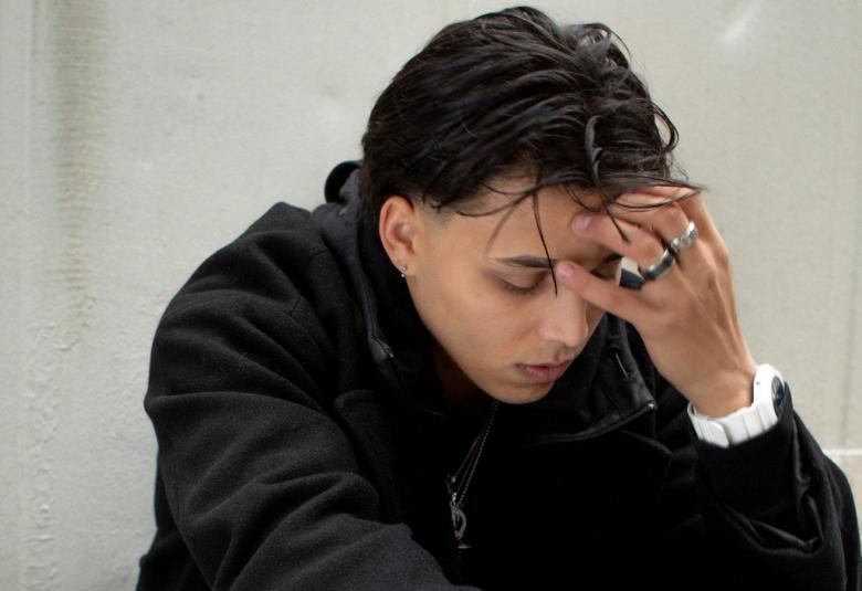 Kuvassa on Averagekidluke istumassa puolivartalokuvassa kuvartuneena. Hän pitää vasemmalla kädellään otsaansa. Tausta on vaalean harmaa.