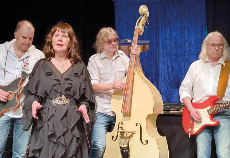 Kuvassa on vasemmalta kitaristi ja hänen vieressään edessään nainen ruskeassa rimpsutetussa mekossa ja keskellä on vaaleaa bassoa soittava mies. Oikealla on kitaraa soittava mies valkoisessa paidassa ja farkuissa.