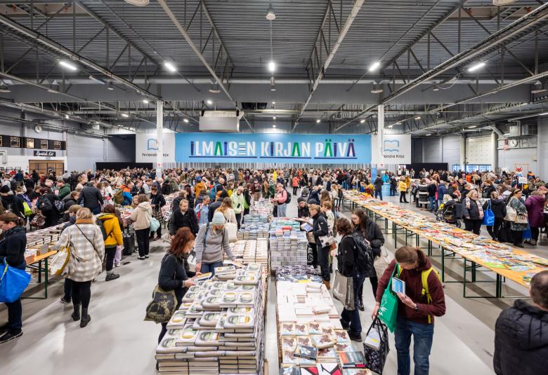 Kuvassa on isossa hallissa useita pitkiä pöytiä täynnä kirjoja ja niitä katselemassa on paljon ihmisiä. Takana on iso sininen lakana, jossa on valkoisin kirjaimin Ilmaiskirjapäivä.