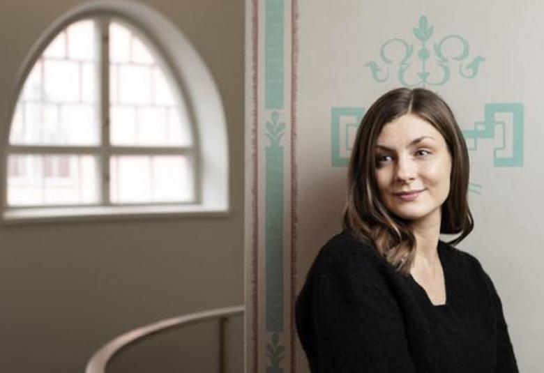 Kuvassa on oikealla Iida Turpeinen puolivartalokuvassa mustassa puserossa. Hän on tilassa, jossa on vaalea, turkoosikoristeinen seinämän reunus. Taaempana näkyy kaarevaa ikkunaa.