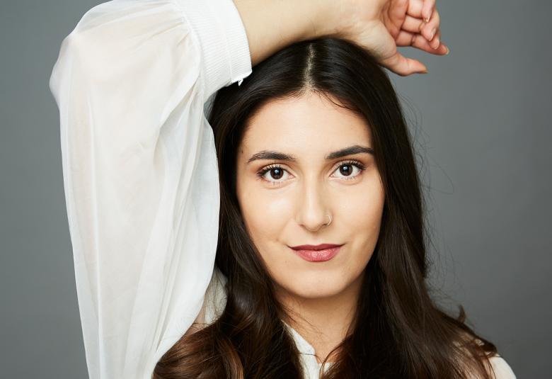 Kuvassa on Iris Candeleria puolivartalokuvassa ja hänellä on pitkt tummat hiukset. Hänellä on valkoinen pitkähihainen pusero ja hän pitää oikeaa kättään päälaellaan. Tausta on harmaa.