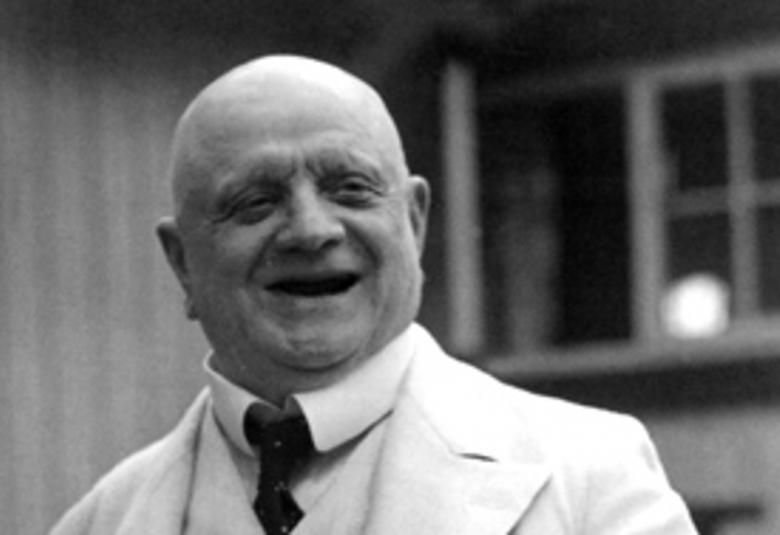 Kuvassa on Jean Sibelius kaljupaisenä puolivartalokuvassa ja hänellä on valkoinen takki ja paita sekä m usta solmio. Taustalla näkyy talon seinää ja ikkunaa. Kuva on musta-valkoinen.