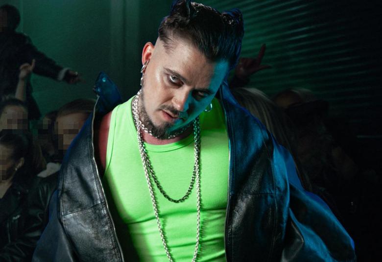 Kuvassa on Käärijä puolivartalokuvassa. Hänellä on hihaton vaalean vihreä t-paita ja musta nahkatakki olkapäillään kevyesti. Taustalla on tummahkoa ja alempana näkyy yleisön kasvoja.