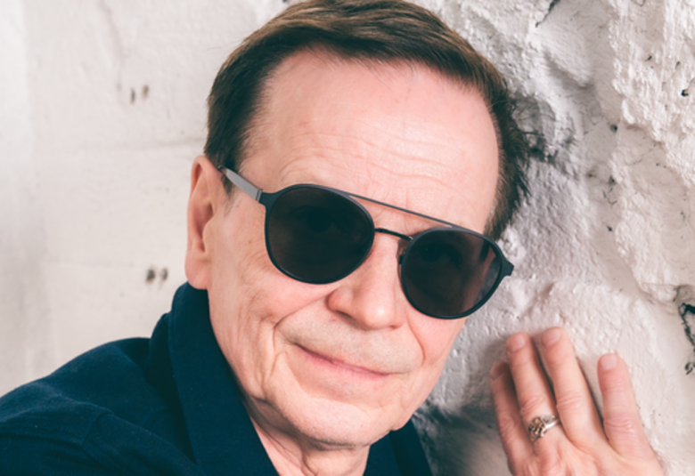 Kuvassa on Kake Randelinin kasvot.  Hänellä on aurinkolasit päässään ja hän pitää oikeaa kättään vaaleassa tiilisessä seinässä.  