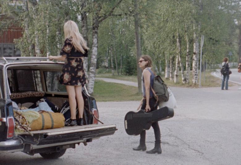 Kuvassa on vasemmalla auton takakontti auki ja sen päällä seisoo reunalla selin Alina Tomnikov. Taustalla on koivumetsää ja asfaltilla oikealla kävelee poispäin nuori mies.